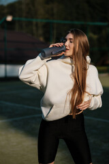 Fototapeta premium Young lovely long hair girl athlete sensually drinking water from plastic bottle on tennis court. Fashion and sport concept.