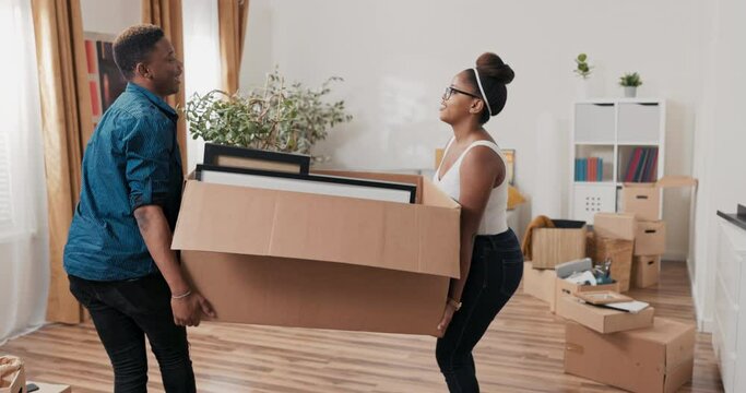 A young couple in love bring heavy boxes of packed belongings picture frames into their purchased rented home during a move, place a cardboard box on the floor and high-five