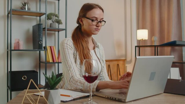 Young woman freelancer drinking wine from glass while working typing on laptop computer sitting at desk at home office. Focused female girl businesswoman working from distant workplace, remote job