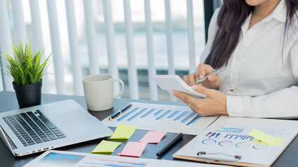 An Asian accountant's hand is pressing a calculator and simultaneously analyzing financial graphs and a laptop next to it while still holding a pen in his finger.