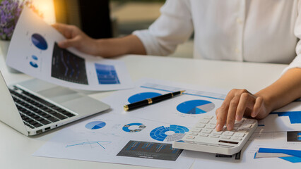 An Asian accountant's hand is pressing a calculator and simultaneously analyzing financial graphs and a laptop next to it while still holding a pen in his finger.