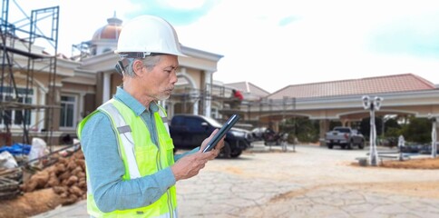 A senior Asian architect wearing a helmet and safety vest uses a tablet to frame a construction site.