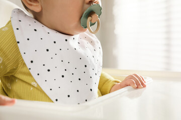 Cute little baby wearing bib in highchair indoors, closeup