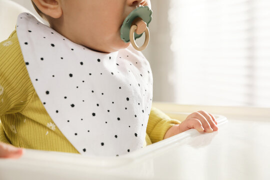 Cute little baby wearing bib in highchair indoors, closeup
