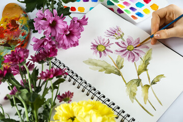 Woman painting chrysanthemums in sketchbook and flowers at white table, closeup