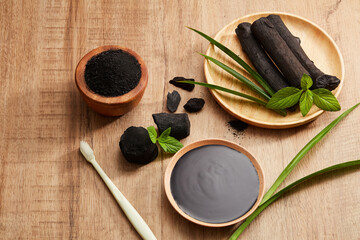 Flatlay of bamboo activated charcoal and charcoal mask and scrub , toothbrush decorate in wooden dish with green leaf in wooden background 