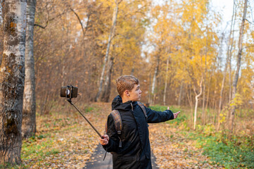 A teenage boy in a black jacket and a monopod in his hand shoots video on a smartphone. Boy blogger. Walk through the autumn park. Close-up.