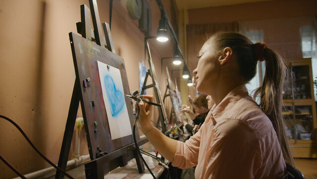Woman artist learns to paint with airbrush with acrylic dye, paper and easel. Indoors. Concept modern art, airbrushing, aerography, draw spraying picture create sprayer drawing graffiti aerosol spray