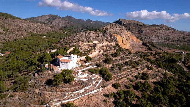 Aerial 4K footage of a small white hermitage among trees on top of a hill next to a castle ruins_Ermita La Magdalena de Castello