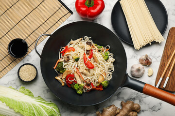 Stir fried noodles with chicken and vegetables in wok on white marble table, flat lay