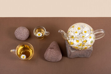 Chamomile tea. Cups of herbal tea, transparent teapot with camomile flowers and stones on brown background. Calming drink concept. Trendy still life