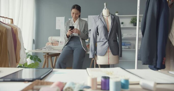 Cheerful Asia Businesswoman Fashion Designer With Suit Collection Smile Sitting On Table Use Smartphone Chat With Customer Confirm Online Purchase Order In Studio Workplace. Start Up Online Business.