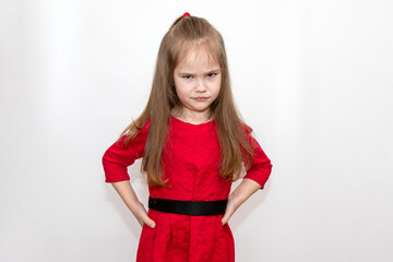 Portrait of a girl in a red dress with long hair on a white background. The child stands frowning, dissatisfied with his hands on his belt.