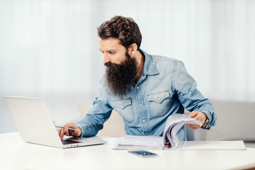 A bearded freelancer going trough paperwork and typing on his laptop at home.