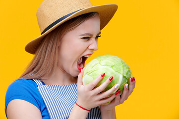 Young woman farmer wearing straw hat and apron biting cabbage on yellow background