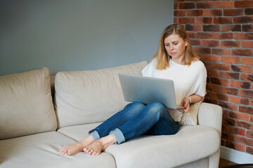 Thoughtful woman holding laptop on her lap, thinking over ideas or tasks, sitting on the sofa at home, dreamy woman, in casual clothes, looking thoughtfully, lost in thought, waiting for a message