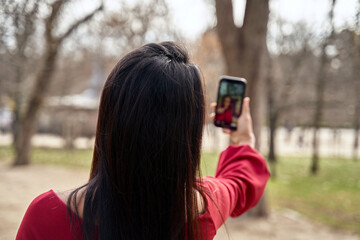 Back view of anonymous female with black hair taking self portrait on smartphone while standing in park with high trees