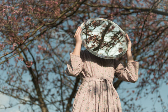 girl in classic floral dress under cherry blossom tree holding large round mirror