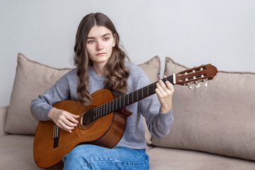 Fototapeta premium A teenager with long dark hair is sitting at home on the couch and playing the guitar.