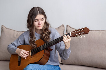 Fototapeta premium A teenager with long dark hair is sitting at home on the couch and playing the guitar.