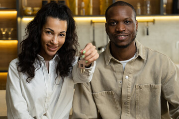 Happy couple shows key on ring from apartment in kitchen. Mixed race woman and African American man smile after successful bargain. Young owners of new home closeup