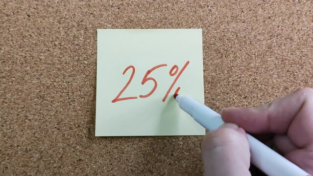 The phrase 25% on a yellow paper sticker. Female hand writing with a felt-tip pen. A red marker in a woman's palm. Sticker on a cork board close-up.