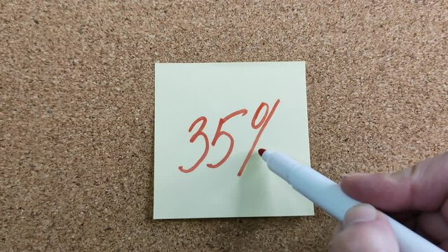 Writing 35% on a yellow paper sticker. Female hand drawing with a felt-tip pen. A red marker in a woman's palm. Sticker on a cork board close-up.