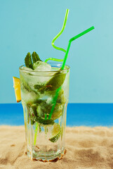 Green cocktail with a straw on the sand. Alcoholic mojito with ice, lime and mint on the beach.