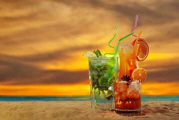 Orange and green cocktails on the beach. Alcoholic drink with ice, orange, mint and lime close up. Sunset, sea and sky in the background. Copy space and free space for text near the glass.