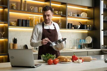Young man in apron prepares dinner following video. Chef cooks dish with vegetables mixing sauce in...