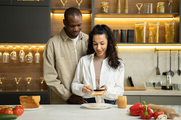 African American man helps beloved mixed race woman spread sauce on bread in modern kitchen. Happy couple hugs cooking dinner against countertop slow motion