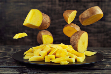 Raw potatoes fall on a black plate. Potatoes fly over a wooden table. Whole and sliced potatoes. Healthy food and farming concept.