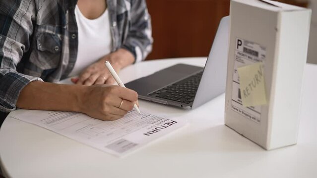 Young female client writing refund and sitting at table with laptop in home room spbd. Closeup view of young african woman fills out document and wants to return order, uses computer and sits at desk