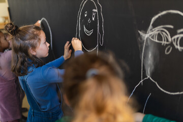 Little girls drawing with chalks on blackboard wall indoors in playroom.