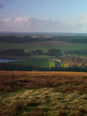 Clouds cast shadows over moorland, wooded hills and a reservoir spillway, all lit up with rich,...