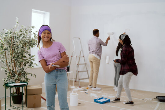 Renovation of apartment, pretty smiling girl in a purple blouse and headband looks at the camera proudly holding paintbrush in hand, in the background the family paints the walls next to the ladder