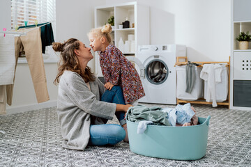 Mother sits on the laundry room floor with daughter cute little girl with blonde hair tied up in a...