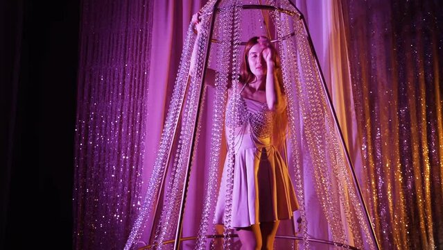 Attractive young woman with long hair dances in shiny silver dress with skirt in large crystal chandelier in multicolored pink light. Beautiful decorations from beads in photo studio