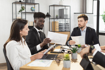Multiracial company workers in formal wear examining financial report during meeting at boardroom. Confident men and women having discussion while sitting at office desk.