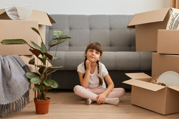 Image of pensive little dark haired girl wearing casual style clothing sitting on floor near sofa...