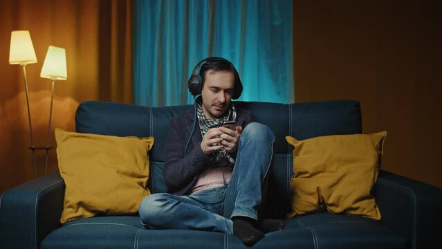  Caucasian man in the headphones listening to the music on the smartphone through headphones in the living room.