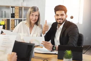 Young indian man and caucasian senior woman smiling and looking at camera during paperwork at office and showing thumb up. Two company workers in formal wear cooperating together.