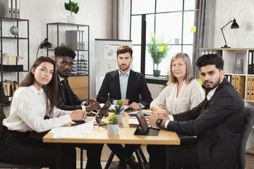 Team of five multiracial business people in formal clothes sitting together at office desk, smiling and looking at camera. Conference meeting of diverse colleagues.