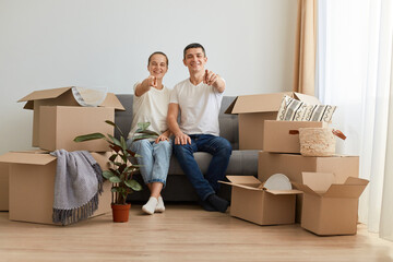 Indoor shot of excited happy young couple sitting on sofa surrounded with carton boxes with personal belongings, pointing to camera, choosing you for buying new apartment, mortgage.