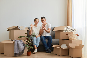 Horizontal shot of smiling young family buying new flat and posing surrounded with boxes with personal pile, sitting on sofa and having video call, waving hands to phone camera.
