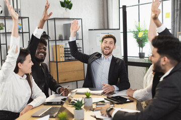 Happy multiracial people in formal clothes sitting together at desk and raising their hands during meeting. Group of business colleagues voting with one voice for common working project.