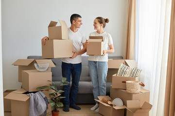 Portrait of happy family, woman and man wearing white t shirts standing with cardboard boxes, relocating to a new house, unpacking personal belongings, looking at each other with love, real estate.