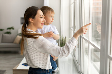 Happy young mother holding baby son on hands, standing by window and showing him world outside