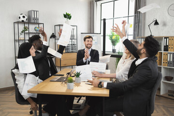 Team of happy diverse business partners sitting at desk and throwing up documents during successful meeting at office. Agreement, cooperation and people concept.