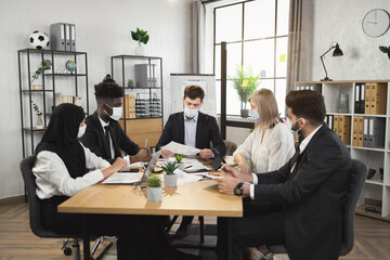 Multi ethnic financial specialists in formal wear and medical face masks gathering at boardroom for conference meeting. Brainstorming of male and female business people.